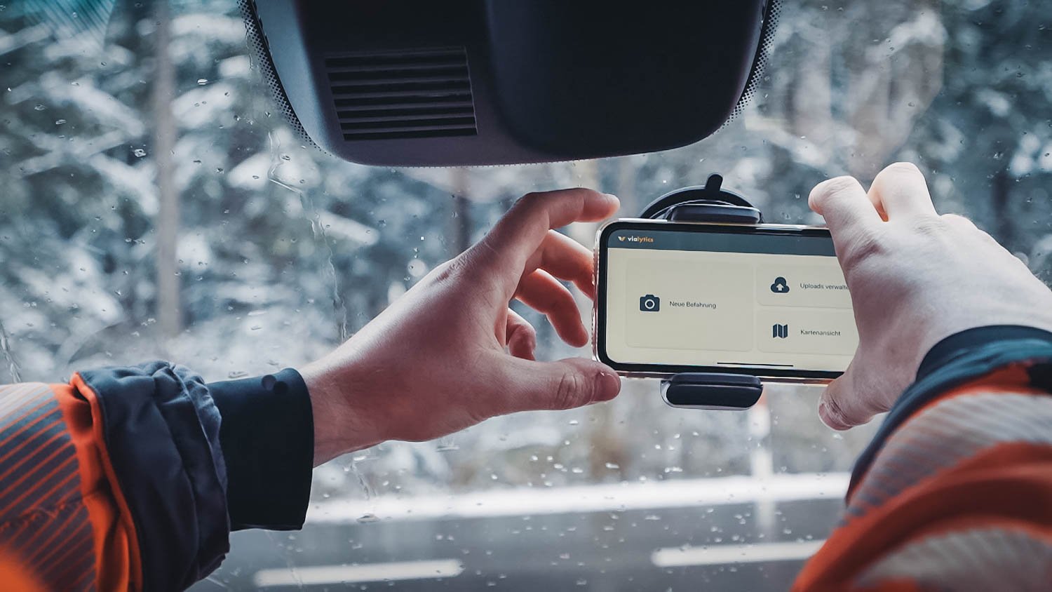 Eine Person vom Straßenbau befestigt ein Handy in einer Halterung an der Frontscheibe seines Räumungsfahrzeuges. Im Hintergrund ist unscharf Schnee zu erkennen