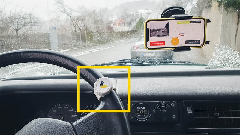 Zu sehen ist ein Auto von innen. Am Lenkrad ist der vialytics Bluetooth-Knopf angebracht. An der Windschutzscheibe ist das Handy mit dem System zur Straßenzustandserfassung angebracht