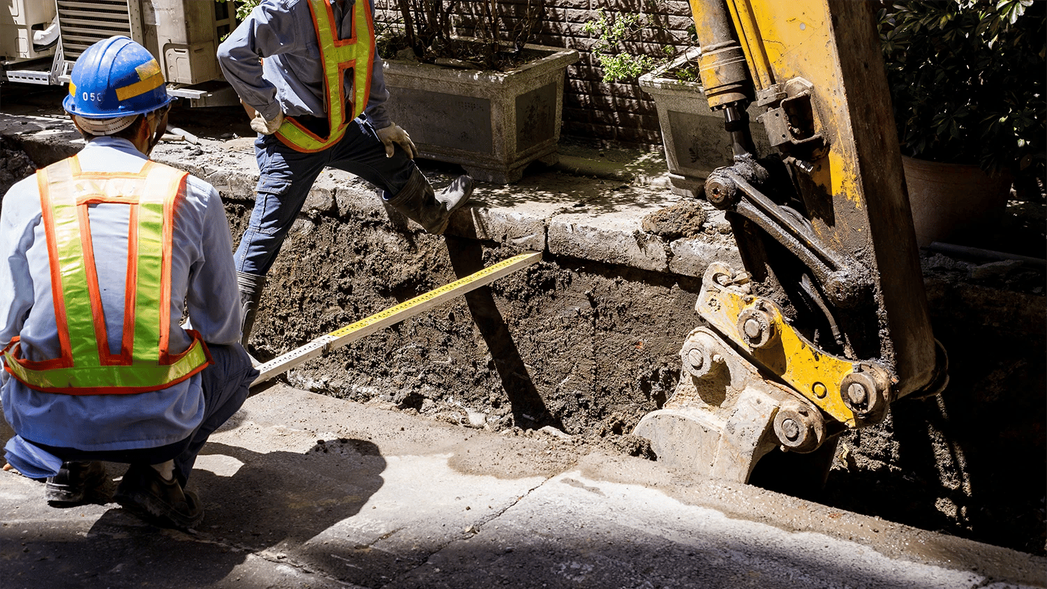 Ein Bagger gräbt eine Straße auf und zwei Bauarbeiter messen das Loch aus