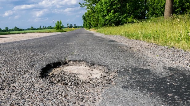 Landstraße bei Tag, Bild auf Höhe der Asphaltdecke zeigt ein großes Schlagloch im Vordergrund