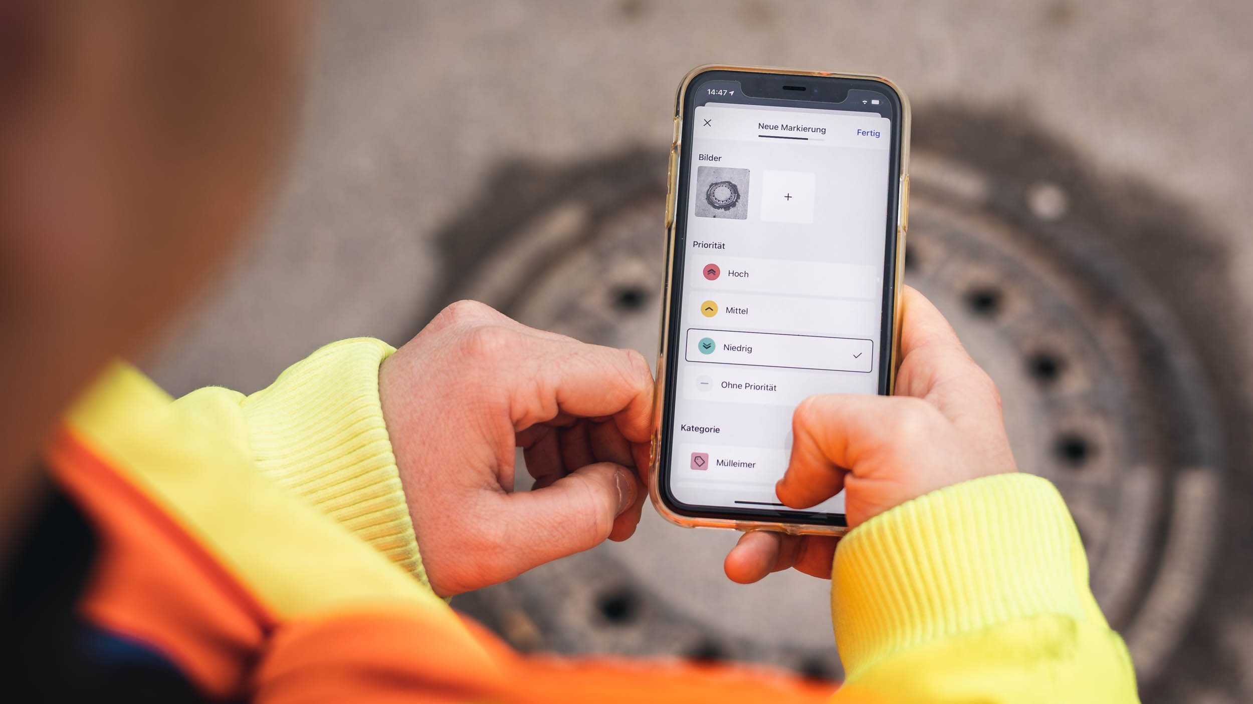 Ein Mitarbeiter des Bauhofs hält ein Handy in der Hand und hat das Markierungstool geöffnet. Im Hintergrund ist eine Schachtabdeckung zu sehen, eine Aufgabe der Verkehrssicherungspflicht