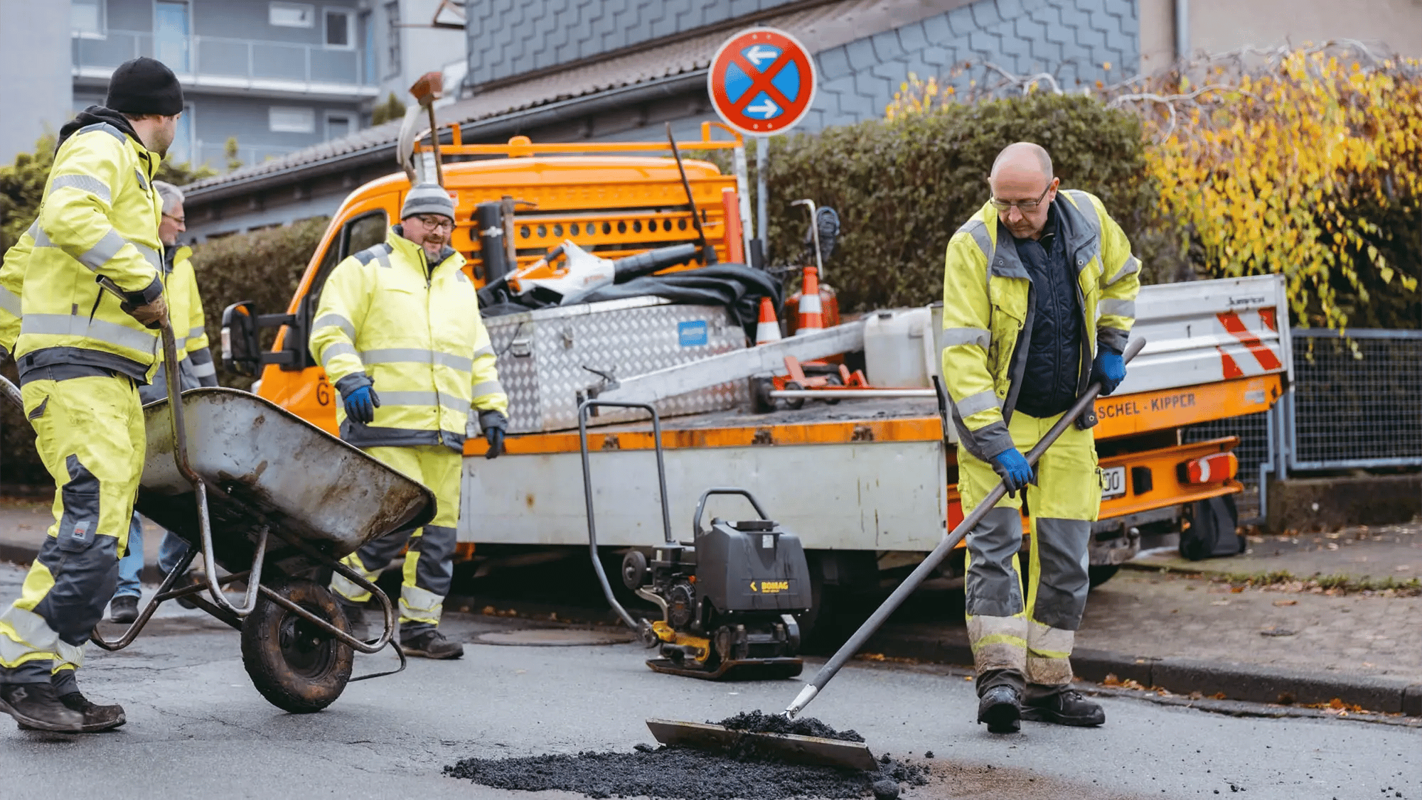 3 männliche Personen reparieren einen Straßenschaden in Goslar. Ein Baufahrzeug ist im Hintergrund, mehrere Maschinen kommen zum Einsatz.