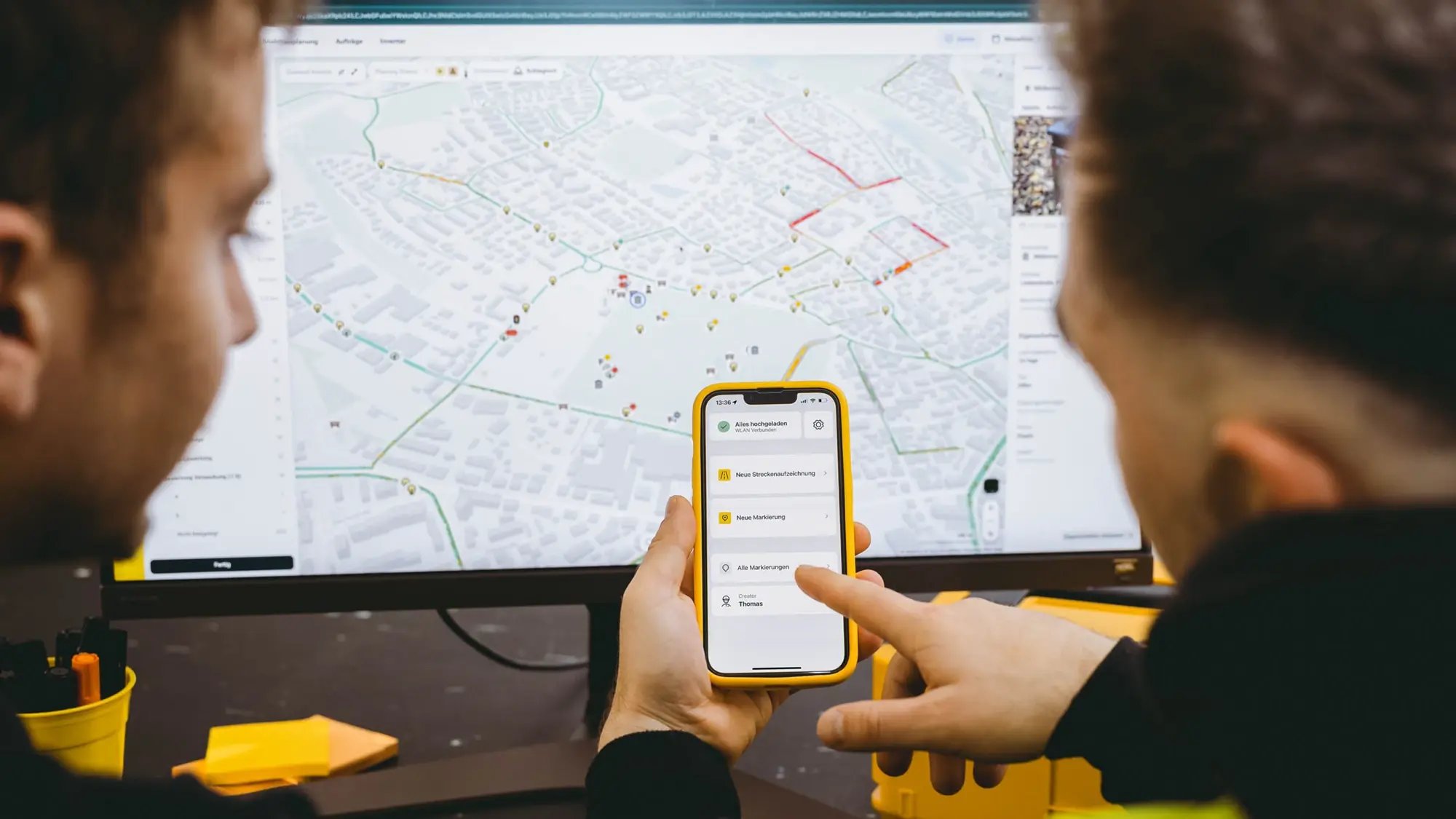 2 Personen sitzen vor einem Monitor mit dem vialytics Webssystem und unterhalten sich und zeigen auf ein Samrtphone mit der vialytics App
