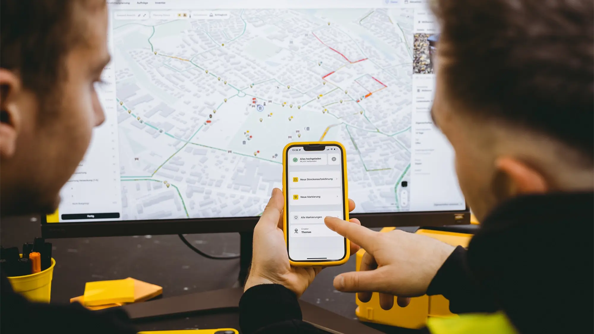 Zwei personen vor einem Monitor mit vialytics System, schauen auf ein Smartphone und zeigen auf den Screen.